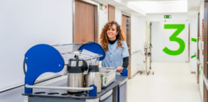 a woman standing in a room with a blue and white counter. Catering Assistant, pushing tea trolley in hospital corridor. 3Q Recruitment.