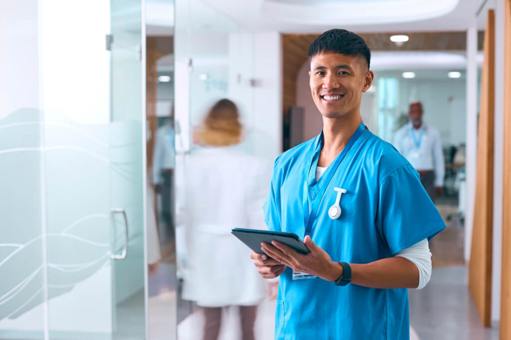 How To Advance Your Career As A Staff Nurse 2 Smiling male nurse with busy hospital ward background. Staff Nurse.